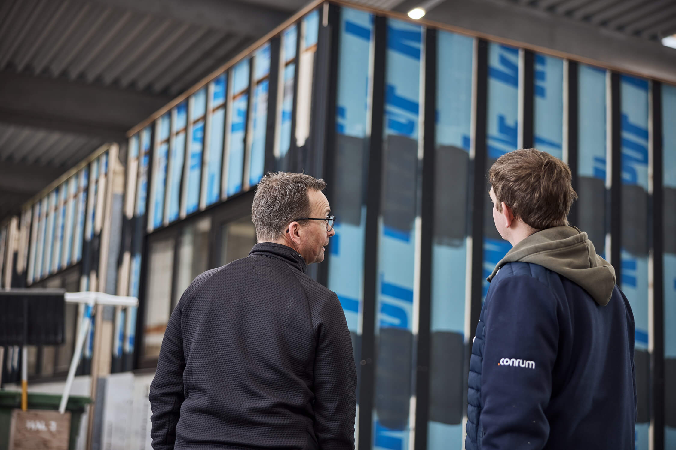 Two men in jackets talking outside a modern glass-front building with blue banners.